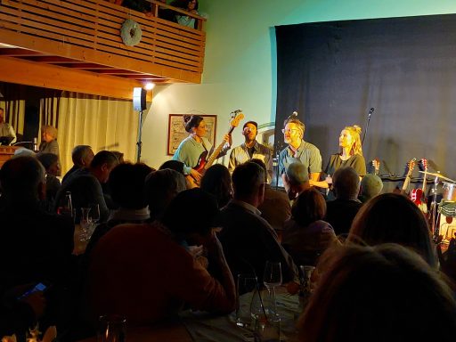 Eine Band spielt live vor einem begeisterten Publikum in einem gut gefüllten Raum.